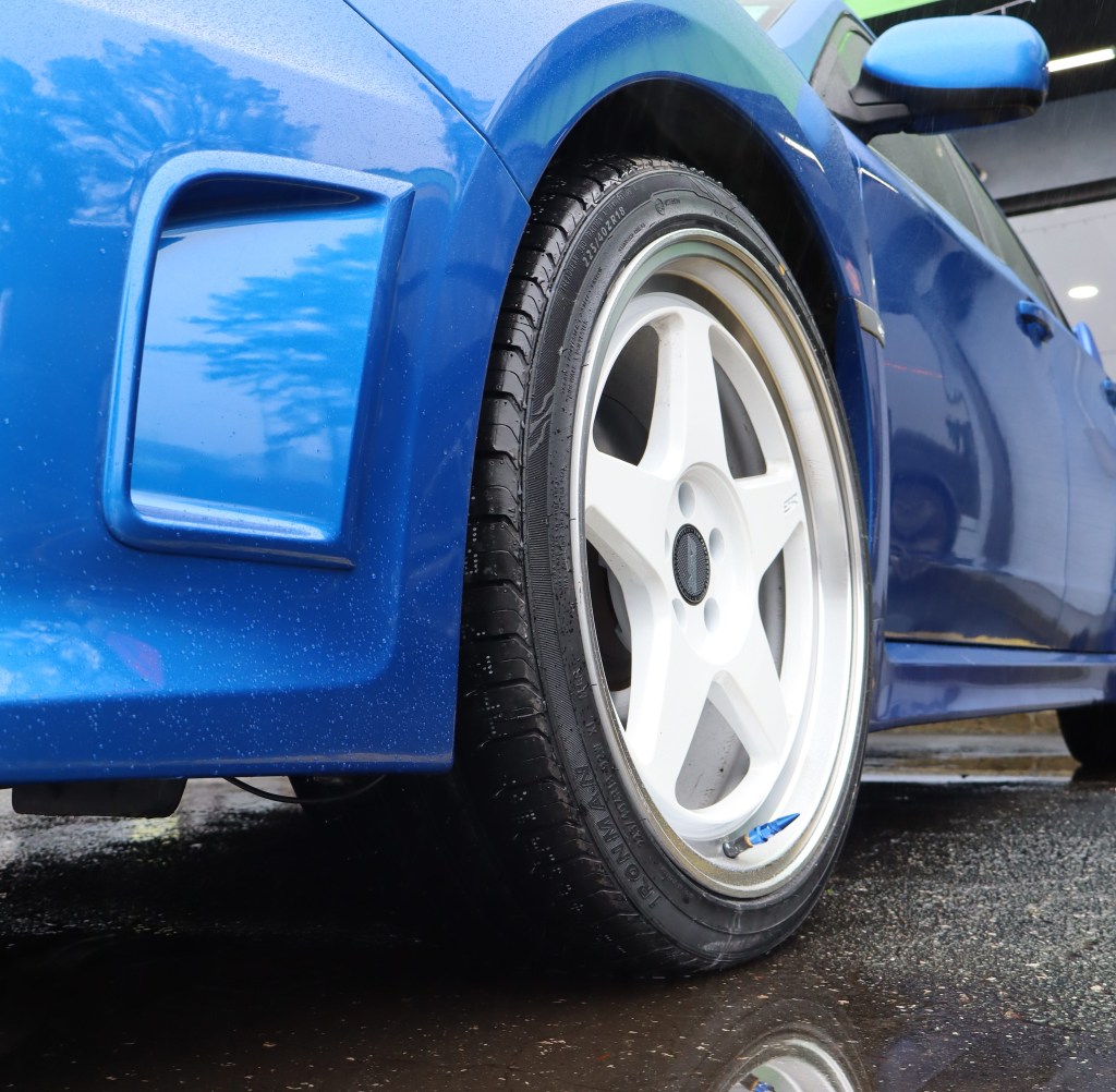 A blue car with white sport wheels with a reflection on the water